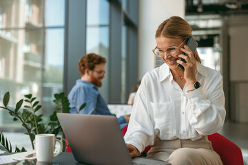 Wall Mural - Businesswoman talking on phone with client while working on laptop in office . High quality photo