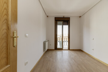 Empty room in a house with white painted walls, wooden floors, white aluminum radiators and brown aluminum sliding doors leading to a small balcony