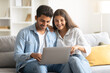 © Prostock-studio - Young Indian couple browsing on laptop, sitting on couch at home