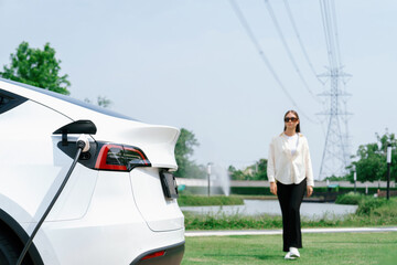 Young woman recharge EV car battery at charging station connected to power grid tower electrical industrial facility as electrical industry for eco friendly vehicle utilization. Expedient