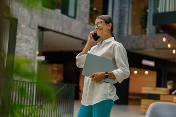 Business woman talking phone with client while walking in office hall with laptop