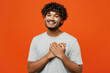 Young cheerful smiling fun happy grateful Indian man he wears t-shirt casual clothes put folded hands on heart look camera isolated on orange red color background studio portrait. Lifestyle concept