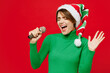 © ViDi Studio - Young singer woman in green turtleneck Santa hat posing sing song in microphone ar karaoke club spread hand isolated on plain red background Happy New Year 2024 celebration Christmas holiday concept