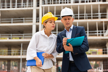 two confident engineers in hard hats discussing blueprint while standing at construction site