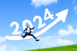 © Creativa Images - Business woman jumping over arrow sign with 2024 on the grass