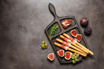 Wall Mural - Bread sticks grissini with prosciutto ham , blue cheese, figs, grape on dark rustic background, closeup. Italian antipasti . Top view, flat lay, vertical