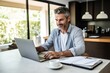 Smiling elegant businessman holding documents and looking at camera while working from home. Freelancer working from home sitting at desk with laptop. Self employed business person working from home.