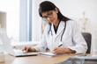 © sofiko14 - Side view of indian female doctor using portable computer while sitting at office desk in health center interior. Efficient mature general practitioner checking patient records via gadget.
