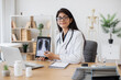 © sofiko14 - Concentrated female doctor talking on video call while showing result of examination of patient with tuberculosis. Indian lady consulting person using laptop and tablet in spacious clinic room.
