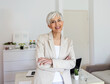 © Jelena Stanojkovic - Confident female looking at camera while standing in home office. Cropped portrait of an attractive senior businesswoman sitting at the desk with her arms crossed while working from home.