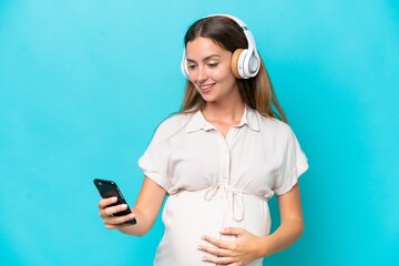 Wall Mural - Young caucasian woman isolated on blue background pregnant and listening music with a mobile