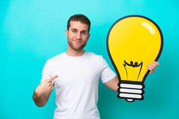Wall Mural - Young caucasian man isolated on blue background holding a bulb icon and pointing it