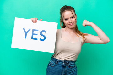 Wall Mural - Young caucasian woman isolated on green background holding a placard with text YES doing strong gesture