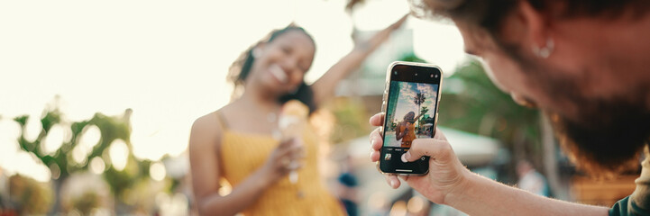 Wall Mural - Close-up of a man taking a picture of a woman on a mobile phone. Closeup, smiling woman eating ice cream and filming it with smartphone on urban city background. Backlight