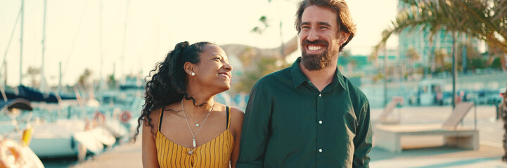 Wall Mural - Close-up of young happy man and woman walking along the promenade holding hands and joyfully chatting on yacht background. Closeup, joyful date of a young interracial couple.