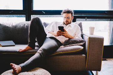 Poster - Serious caucasian man in spectacles dialing number on mobile phone lying on sofa in home interior,