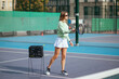Positive beautiful woman tennis coach serving balls from basket to student on tennis court and smiling