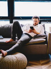 Poster - Serious caucasian man in spectacles dialing number on mobile phone lying on sofa in home interior,