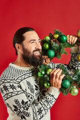 Canvas Print - happy bearded man in winter sweater holding decorated wreath with baubles on red backdrop, Christmas