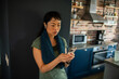 © Marko Geber - Asian woman taking a break in her kitchen checking her phone