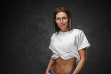 Alluring woman in glasses showcases her sculpted abs in a white crop-top against a dark backdrop