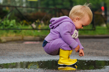 Wall Mural - Saint Petersburg, Russia - 18 July 2023. Baby kids toddler in a jumpsuit of gap company  in yellow boots walks and splashes puddles. Cute happy child in colorful clothes jumping into puddle, splashing