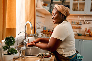 Hygiene on kitchen. Profile of plus size lady wearing headband and apron cleansing her hands in sink before culinary activities. Happy african homemaker getting ready for home-based cooking process.