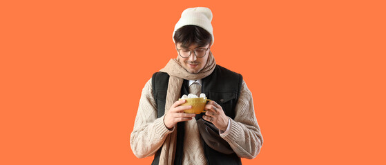 Wall Mural - Stylish young man in winter clothes and with cup of hot chocolate on orange background