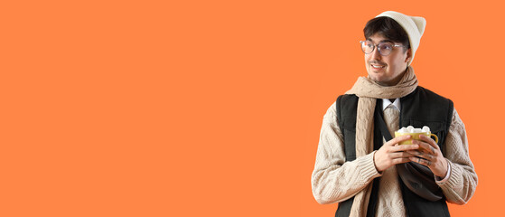 Wall Mural - Stylish young man in winter clothes and with cup of hot chocolate on orange background with space for text