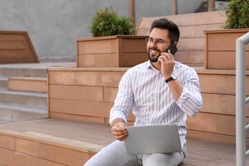 Sticker - Handsome young man talking on smartphone while using laptop on bench outdoors