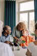 © Maskot - Young female caregiver embracing senior woman sitting with friends at dining table in nursing home