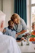 © Maskot - Smiling young female caregiver embracing happy senior men sitting with friends at dining table in nursing home