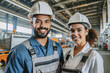 Junger Mann und junge Frau Fachkräfte in Arbeitskleidung und Helmen lächelnd in einer Industriehalle