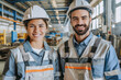 Junger Mann und junge Frau Facharbeiter/Fachkräfte in Arbeitskleidung und Helmen lächelnd in einer Industriehalle