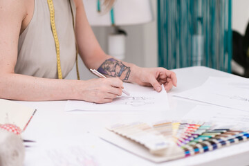 Wall Mural - Lady dressmaker with tattoo draws clothes model on paper at desk in fashion atelier tailor creates pencil sketch of stylish outfit in sewing studio closeup