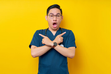 Wall Mural - Shocked young Asian man doctor or nurse wearing a blue uniform and glasses crossed hand and pointing a finger for your choice isolated on yellow background. Healthcare medicine concept