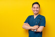 Professional young Asian man doctor or nurse wearing a blue uniform standing confidently while holding stethoscope and crossed hands, smiling friendly at camera isolated on yellow background