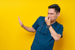 Professional young Asian male doctor or nurse wearing a blue uniform feeling disgusted, holding her nose to avoid smelling foul while showing a stop gesture isolated on yellow background