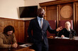 © AnnaStills - Confident attorney in formalwear speaking to prosecution side during trial session while standing against male suspect and mature female judge