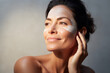 Close-up of middle-aged Caucasian woman touching her face to apply moisturizer. Smiling face of adult brunette lady with daily cream, facial cosmetics. Skin care. Grey background, copy space.