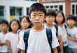 © Gaston - Elementary school student stands in front of school with fellow pupils in the background. Portrait of an asian kid outside the school with classmates in the background