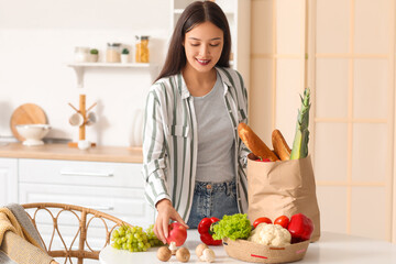 Wall Mural - Young Asian woman unpacking fresh products from market at table in kitchen