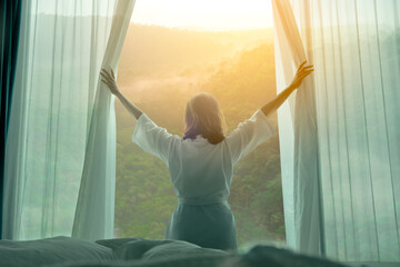 Wall Mural - The woman was at the window in the bedroom. She opened the curtains on the window. In the morning and she looks at the view of mountains and trees at sunrise
