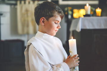 Little kid boy receiving his first holy communion. Happy child holding Christening candle. Tradition in catholic curch. Kid in a white traditional gown in a church near altar.