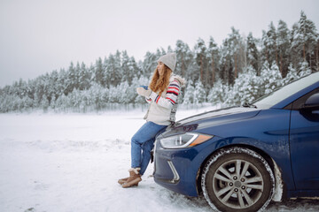 A smiling woman drinks a hot drink from a thermos while standing near her car on a winter snowy road in the forest. The concept of rest, freedom, relaxation, travel.