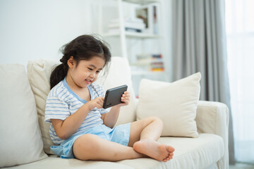 Wall Mural - Asian child girl looking using and touch mobile phone screen on couch sofa. Baby smiling funny time to use mobile phone. Too much screen time. Cute girl watching videos while tv, Internet addiction.