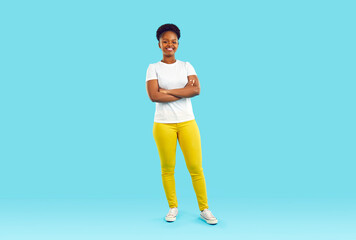 Wall Mural - Full body shot of happy beautiful young African American woman in white Tshirt, yellow pants and comfortable shoes standing with arms folded isolated on blue color background. Casual fashion concept
