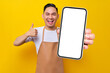 © Bangun Stock Photo - excited young Asian man barista barman employee wearing brown apron work in coffee shop holding mobile phone and showing thumb up sign isolated on yellow background. Small business startup concept