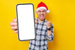 © Bangun Stock Photo - Excited young Asian man in plaid shirt wearing Christmas hat holding mobile phone with blank screen, do winner gesture on yellow background. Christmas and Happy New Year 2023 holiday concept