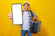 Cheerful young traveler tourist asian man in casual clothes hat holding suitcase and showing blank screen mobile phone isolated on yellow background. Air flight journey concept